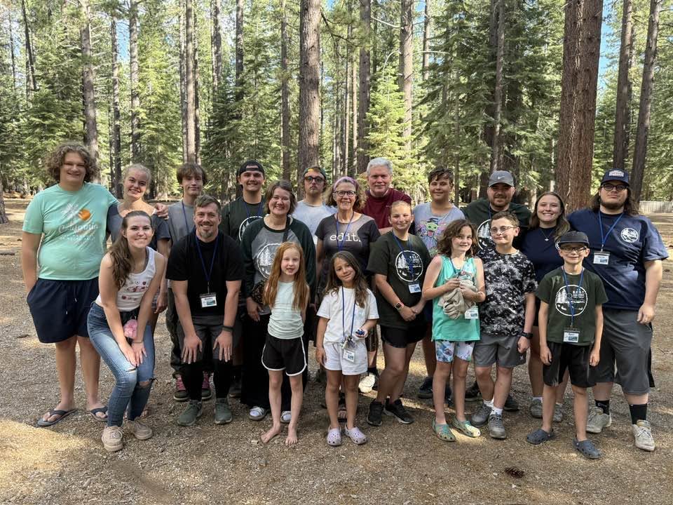 A group photo of 20 people posing together in a forest setting with tall trees, smiling and wearing casual clothing.